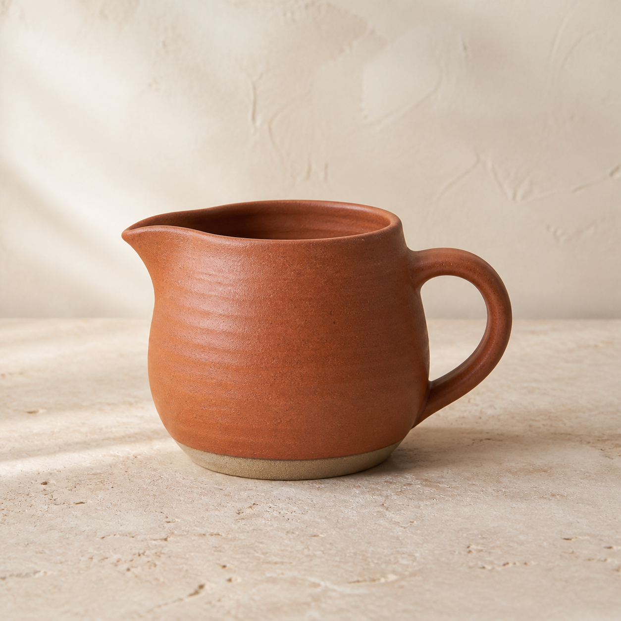 Small terracotta stoneware pourer with a shaped spout, rounded handle and raw clay foot, on a stone bench.